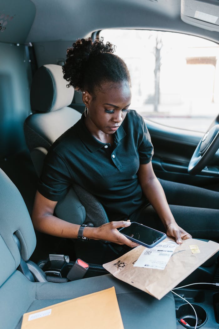 A female courier uses a smartphone to scan a package barcode, seated inside a delivery van.